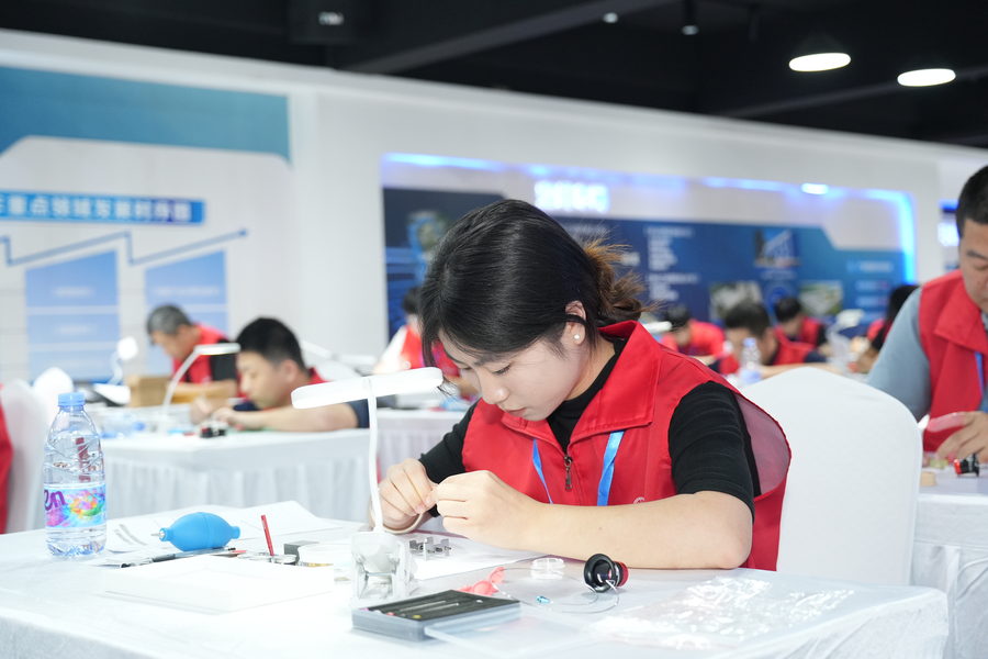 A competitor in a red vest working on a watch movement with tweezers under a bench lamp