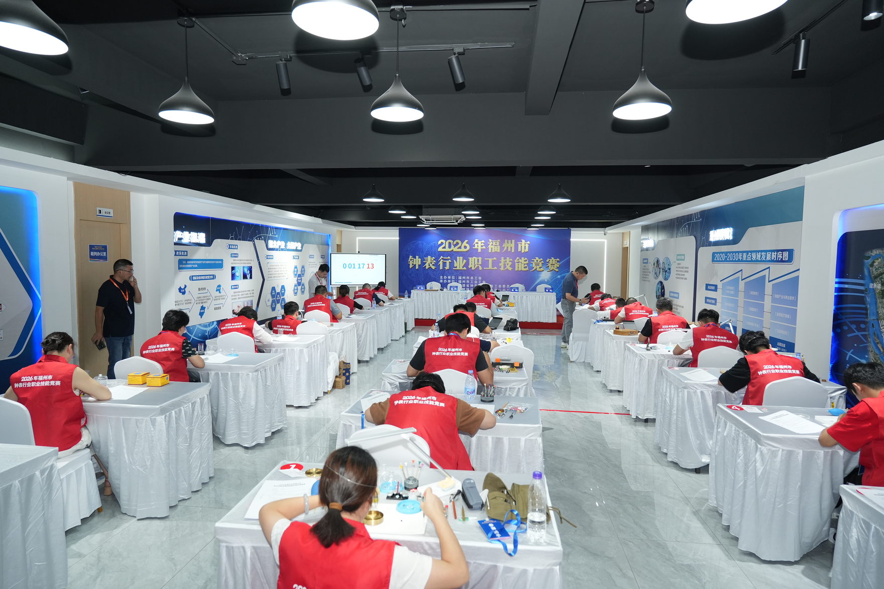 A wide view of the competition hall, with rows of white-draped benches and competitors in red vests