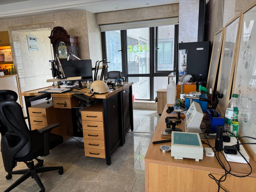 A workstation with a magnifier lamp, drawers and hand tools, and a grandfather clock behind