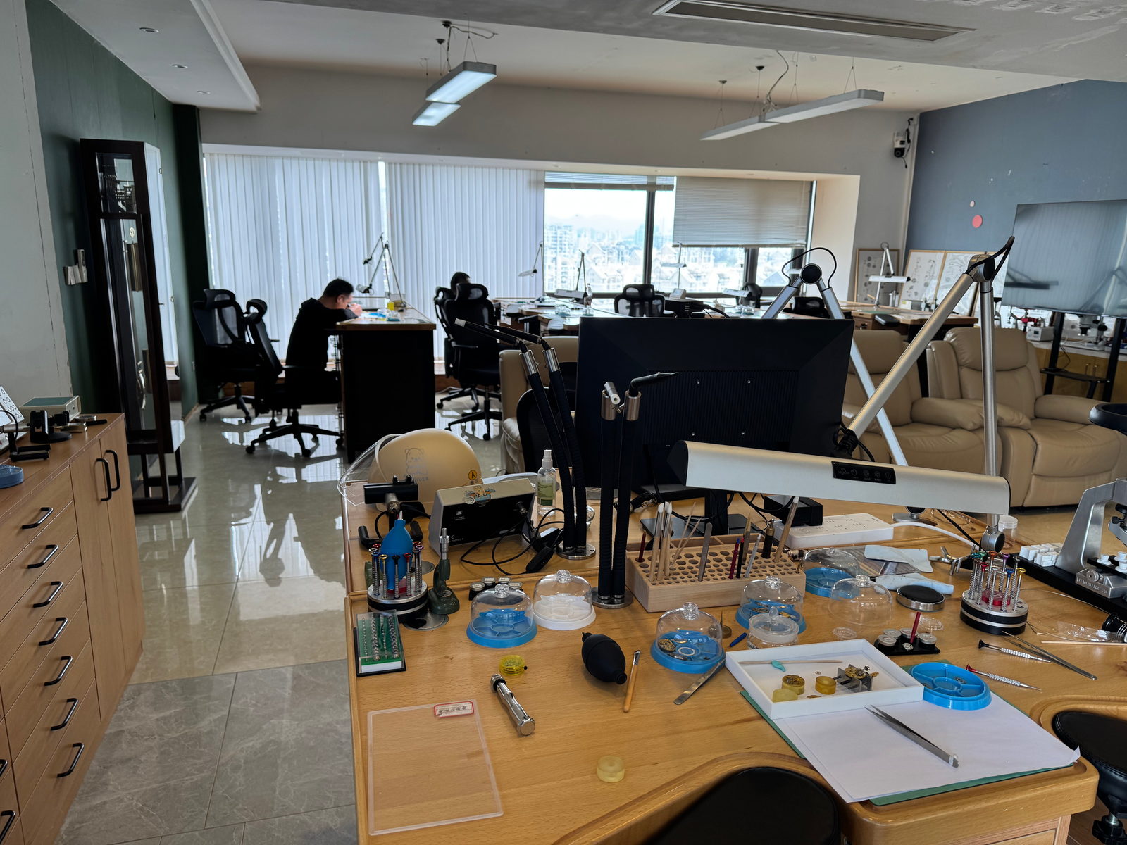 A bench seen from above and laid out with screwdrivers, tweezers, glass dishes and movement trays, with students working at windowside benches behind
