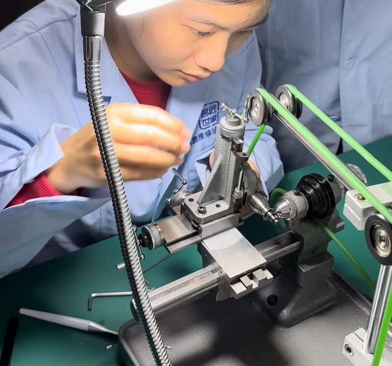 A student in the school's blue workshop coat leaning over a watchmaker's lathe, the green drive belt running past the headstock