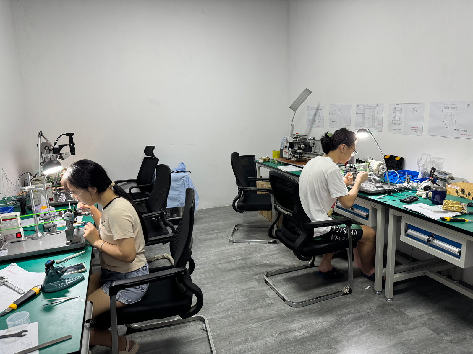 Two students at separate benches in the lathe room, each at a motor-driven lathe under a bench lamp, technical drawings on the back wall